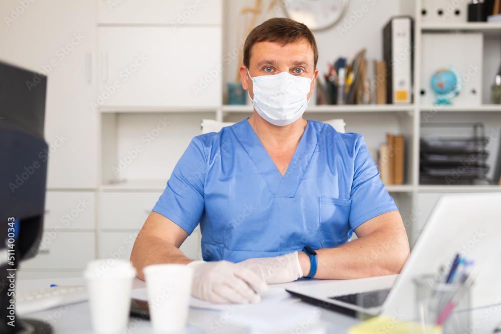 Confident male doctor in blue medical uniform, mask and gloves working on laptop in clinic office