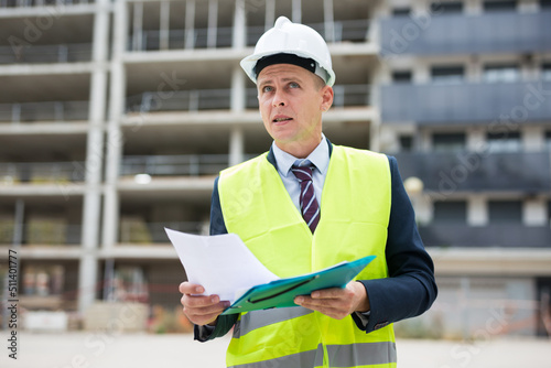 Architect in protective helmet and jacket with folder of documents on the construction site