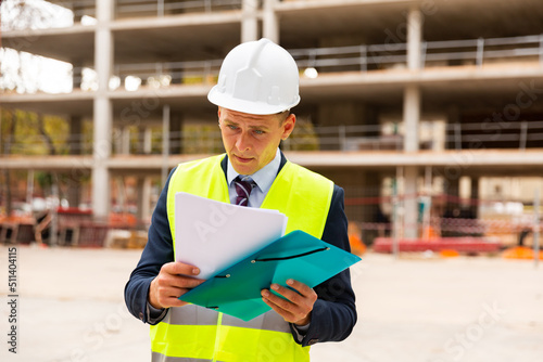 Foreman in protective helmet and vest with folder of documents on the construction site