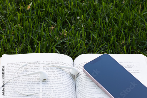 Open bible with mobile phone and headphones laying on the grass with copy space