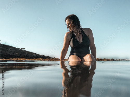 Woman in full piece bathing suit sitting on reef ledge looking into glassy ocean water photo