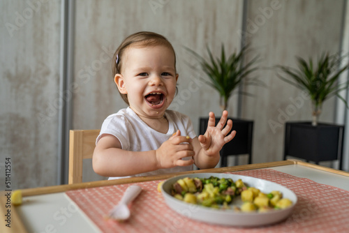 One baby girl small caucasian toddler eating while sit at the table alone at home copy space little child having lunch healthy food meal in room in day happy smile