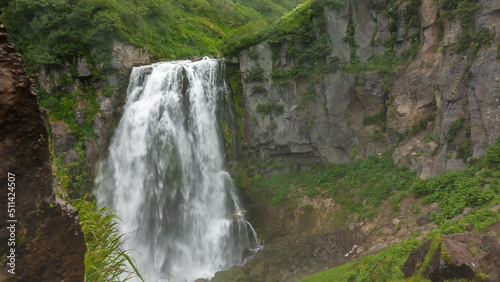 The waterfall flows down from a sheer cliff. Green vegetation on rocky slopes. Kamchatka.