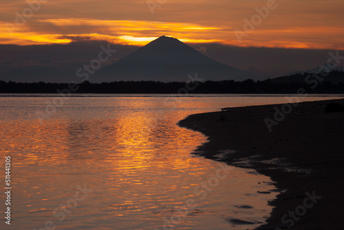 Dramatic golden sea sunset with dark volcano in distance island with colorful orange sky reflection in water, high contrast, black shore. Majestic indonesian landscape in evening, tourism in asia.