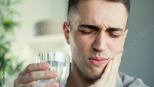 Young man with sensitive teeth and hand holding glass of water	 photo