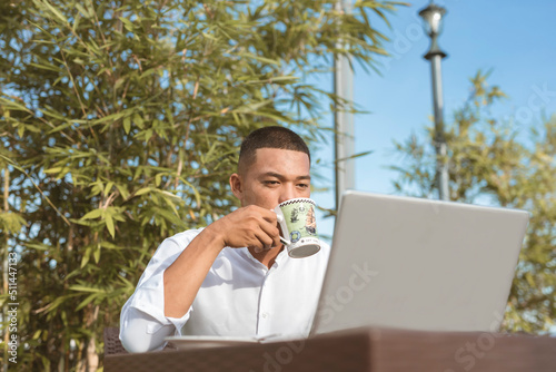 A Filipino male data analyst sips on coffee while working remotely at an outdoor cafe, coffee shop, or restaurant. Modern freelance professional lifestyle. photo