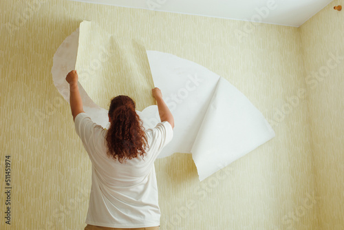 Obraz na plátně Back view of buxom woman with long curly red hair hair wearing white T-shirt, beige trousers, standing on chair, tearing off yellow wallpaper from wall