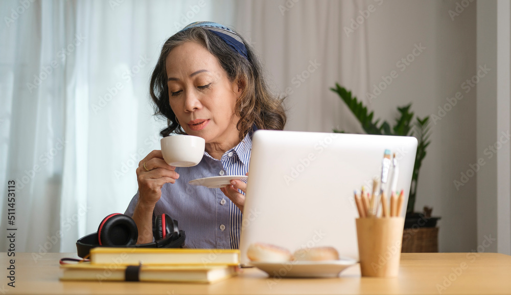 Aged businesswoman drinking hot coffee and using laptop while remote working from home. Aged businesswoman drinking hot coffee and using laptop while remote working from home.