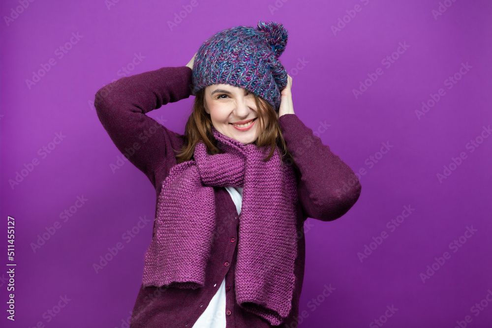 Cute Funny Caucasian Adult Female In Warm Knitted Hat and Violet Scarf Putting Down Hat with Lifted Hands While Having Fun Against Purple Background. Cute Funny Caucasian Adult Female In Warm Knitted Hat and Violet Scarf Putting Down Hat with Lifted Hands While Having Fun Against Purple Background.