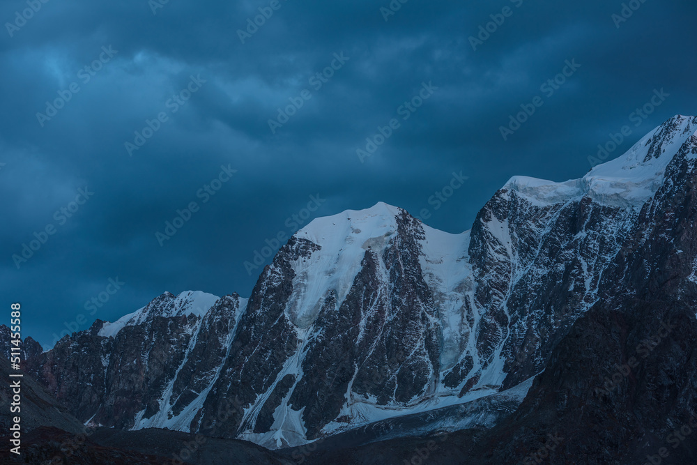 Atmospheric nightly landscape with huge snowy mountain top in dramatic sky. Hanging glacier and cornice on beautiful giant snow mountains in night. High snow-covered mountain range in dusk dim light.