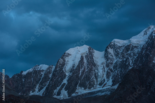 Atmospheric nightly landscape with huge snowy mountain top in dramatic sky. Hanging glacier and cornice on beautiful giant snow mountains in night. High snow-covered mountain range in dusk dim light.