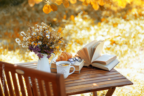 Bouquet of flowers, croissant, cup of tea or coffee, books on table in autumn garden. Rest in garden, reading books, breakfast, vacations in nature concept. Autumn time in garden on backyard