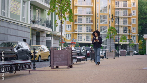 Joyful woman walking texting friends online. Modern residential area district.