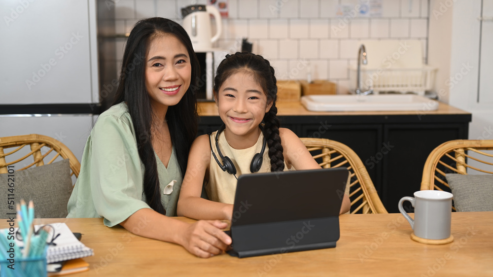 Asian mother and little daughter sitting in kitchen, studying online, doing homework, learning at home, homeschooling concept Asian mother and little daughter sitting in kitchen, studying online, doing homework, learning at home, homeschooling concept