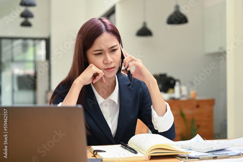 Serious focused female manager sitting in front of laptop and talking on mobile phone, consulting client, making business call at work
