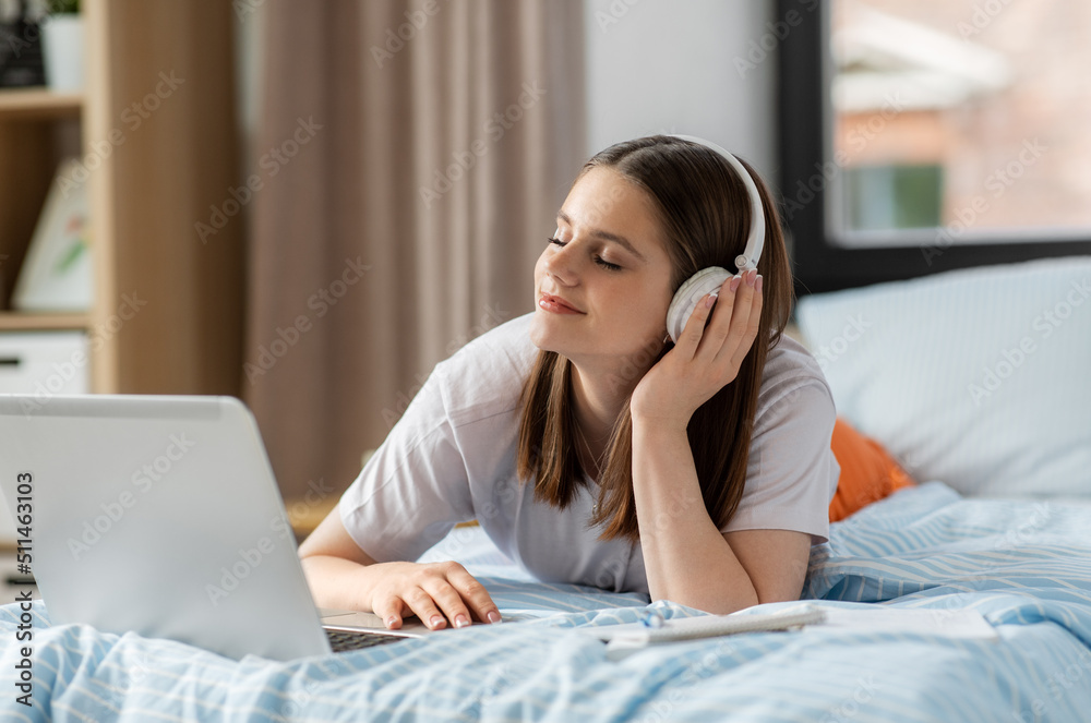 school, online education and e-learning concept - happy smiling teenage student girl in headphones with laptop computer lying on bed and listening to music at home
