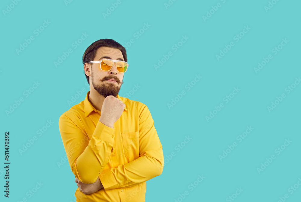 Man with funny expression on his face thinks, imagines or comes up with something interesting. Young caucasian bearded hipster guy holding his chin looking at copy space on light blue background. Man with funny expression on his face thinks, imagines or comes up with something interesting. Young caucasian bearded hipster guy holding his chin looking at copy space on light blue background.