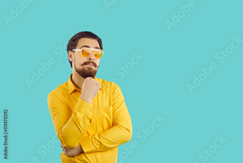 Man with funny expression on his face thinks, imagines or comes up with something interesting. Young caucasian bearded hipster guy holding his chin looking at copy space on light blue background.