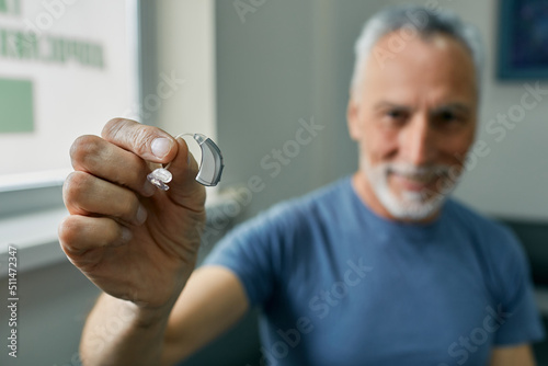Senior man holding BTE hearing aid in hand on foreground, close-up. Treatment of deafness in elderly people photo