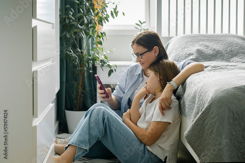 Mother and daughter using smartphone while sitting in berdoom. Woman and pre-teen girl together
