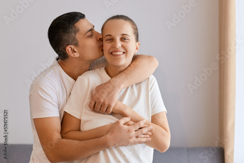 Indoor shot of loving husband cuddling wife from the back while standing in living room, enjoying weekend, celebrating anniversary, expressing love and gentle, smiling. © sementsova321