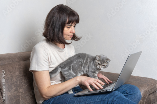 Beautiful middle aged woman with a cutecat works at home on a laptop. Middle-aged woman works remotely. Workplace concept at home with pets. Pets are part of the family photo