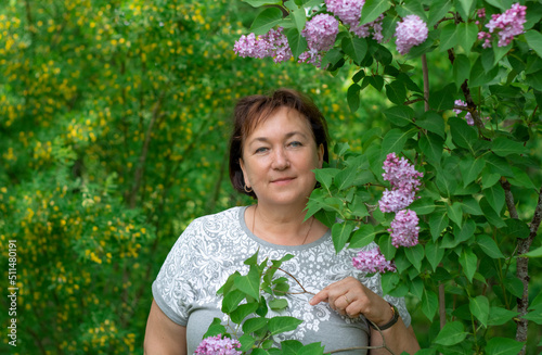 Portrait of an attractive mature woman of 50-60 years old near a lilac bush on a blurry background of flowering acacia. Travel and retirement