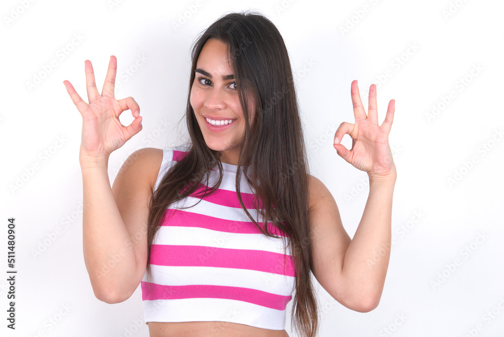 Glad young beautiful brunette woman wearing striped top over white wall shows ok sign with both hands as expresses approval, has cheerful expression, being optimistic.