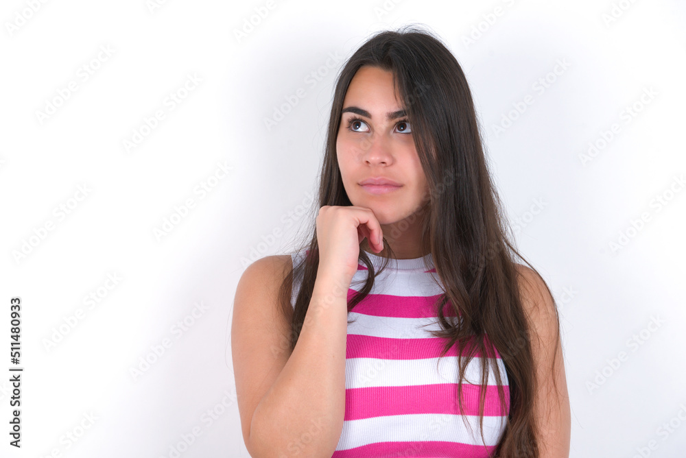 Portrait of thoughtful young beautiful brunette woman wearing striped top over white keeps hand under chin, looks away trying to remember something or listens something with interest. Youth concept. Portrait of thoughtful young beautiful brunette woman wearing striped top over white keeps hand under chin, looks away trying to remember something or listens something with interest. Youth concept.