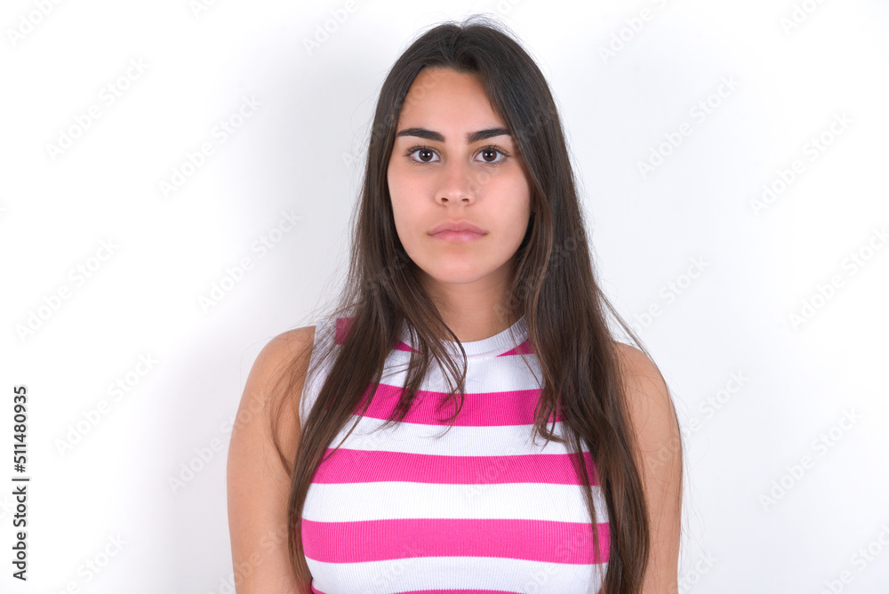 Joyful young beautiful brunette woman wearing striped top over white wall looking to the camera, thinking about something. Both arms down, neutral facial expression. Joyful young beautiful brunette woman wearing striped top over white wall looking to the camera, thinking about something. Both arms down, neutral facial expression.