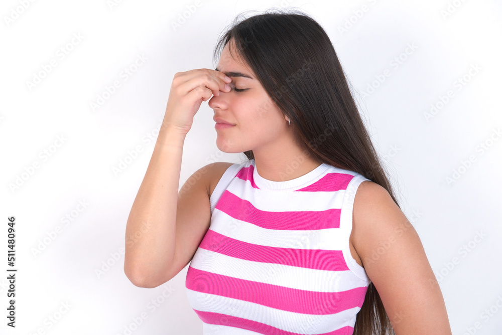 Very upset, young beautiful brunette woman wearing striped top over white wall touching nose between closed eyes, wants to cry, having stressful relationship or having troubles with work Very upset, young beautiful brunette woman wearing striped top over white wall touching nose between closed eyes, wants to cry, having stressful relationship or having troubles with work