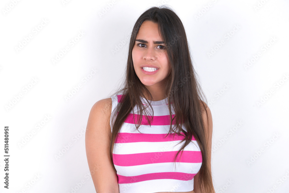 Mad crazy young beautiful brunette woman wearing striped top over white wall clenches teeth angrily, being annoyed with coming noise. Negative feeling concept. Mad crazy young beautiful brunette woman wearing striped top over white wall clenches teeth angrily, being annoyed with coming noise. Negative feeling concept.