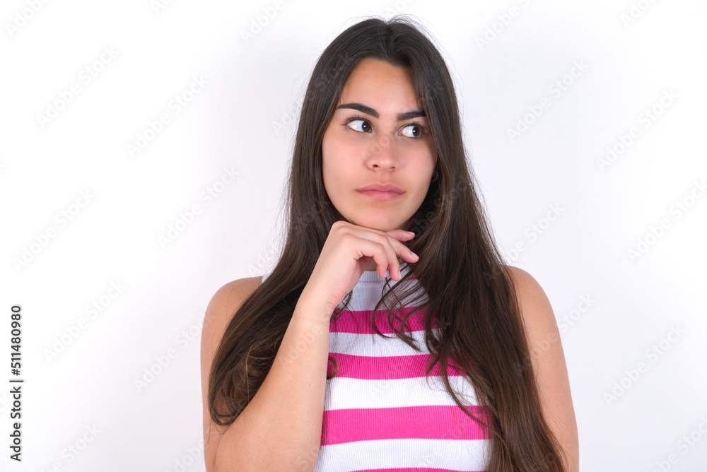 Dreamy young beautiful brunette woman wearing striped top over white wall with pleasant expression, looks sideways, keeps hand under chin, thinks about something pleasant. Dreamy young beautiful brunette woman wearing striped top over white wall with pleasant expression, looks sideways, keeps hand under chin, thinks about something pleasant.