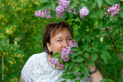 An attractive mature woman of 50-60 years old leans towards a lilac bush on a blurry background of blooming acacia. Travel and retirement