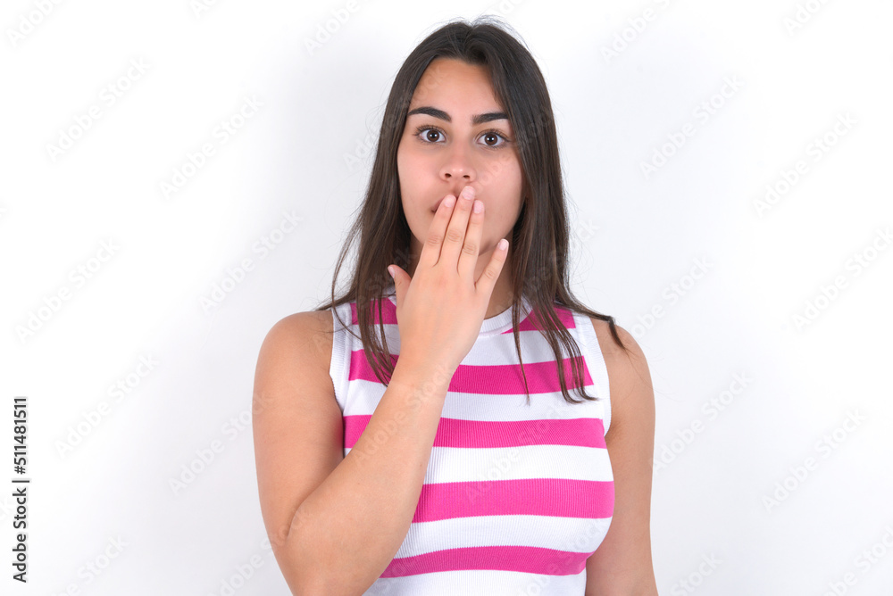 Oh! I think I said it! Close up portrait young beautiful brunette woman wearing striped top over white wall cover open mouth by hand palm, look at camera with big eyes.