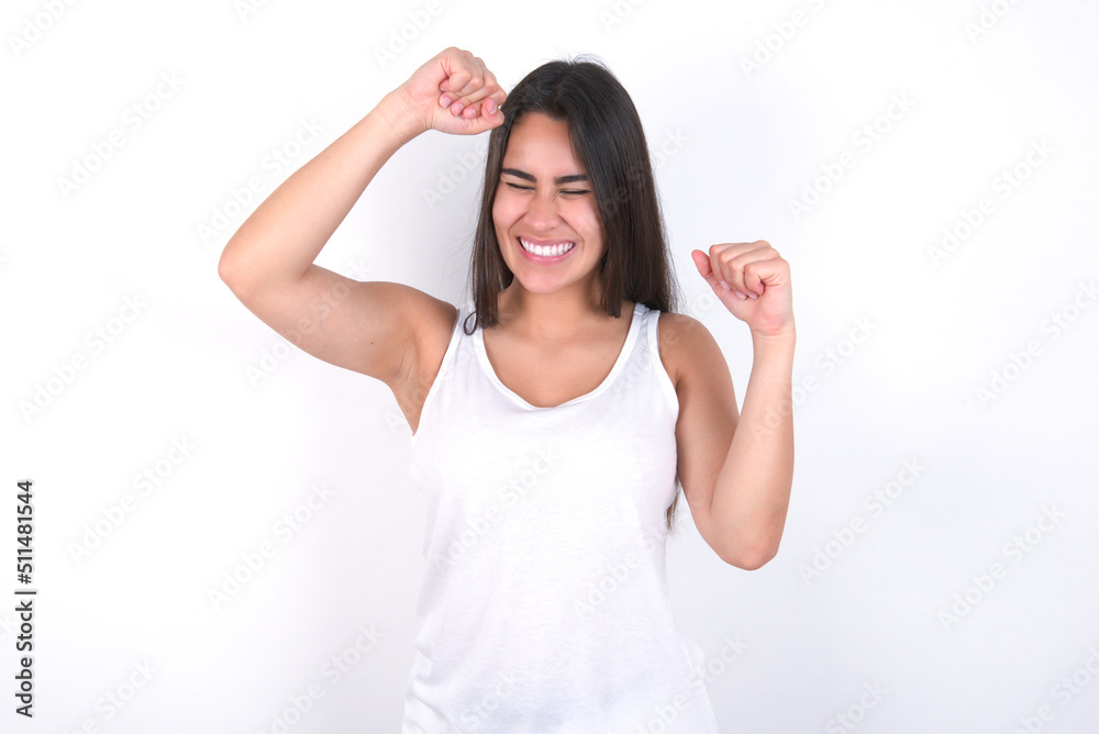 Attractive Young beautiful brunette woman wearing white top over white wall celebrating a victory punching the air with his fists and a beaming toothy smile. Attractive Young beautiful brunette woman wearing white top over white wall celebrating a victory punching the air with his fists and a beaming toothy smile.