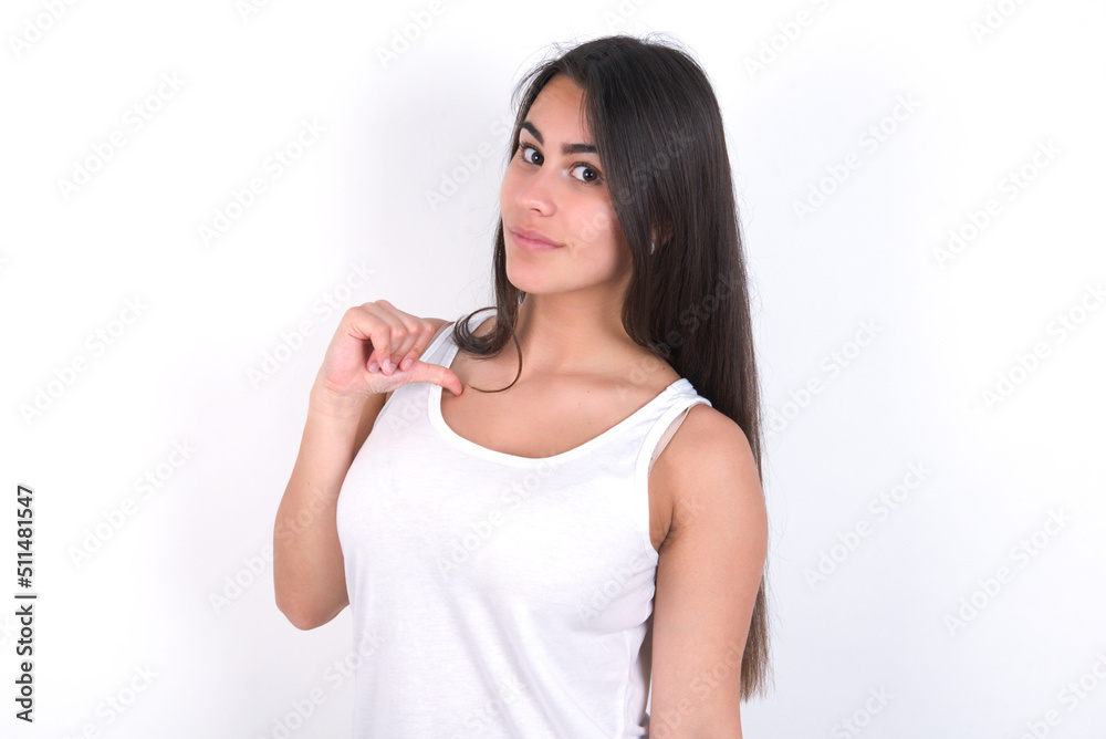 Closeup of cheerful Young beautiful brunette woman wearing white top over white wall looks joyful, satisfied and confident, points at himself with thumb. Closeup of cheerful Young beautiful brunette woman wearing white top over white wall looks joyful, satisfied and confident, points at himself with thumb.