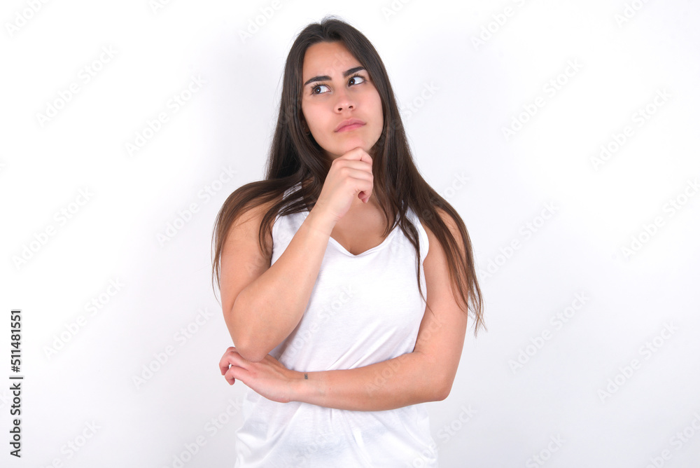 Young beautiful brunette woman wearing white top over white wall with hand under chin and looking sideways with doubtful and skeptical expression, suspect and doubt. Young beautiful brunette woman wearing white top over white wall with hand under chin and looking sideways with doubtful and skeptical expression, suspect and doubt.