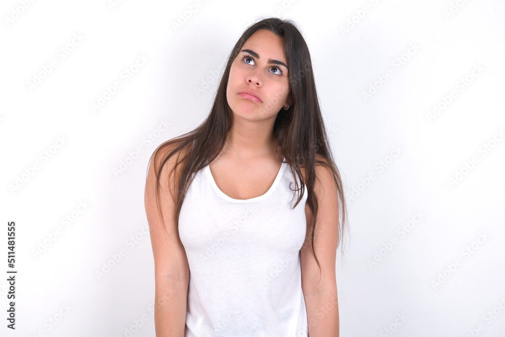 Gloomy, bored Young beautiful brunette woman wearing white top over white wall frowns face looking up, being upset with so much talking hands down, feels tired and wants to leave. Gloomy, bored Young beautiful brunette woman wearing white top over white wall frowns face looking up, being upset with so much talking hands down, feels tired and wants to leave.