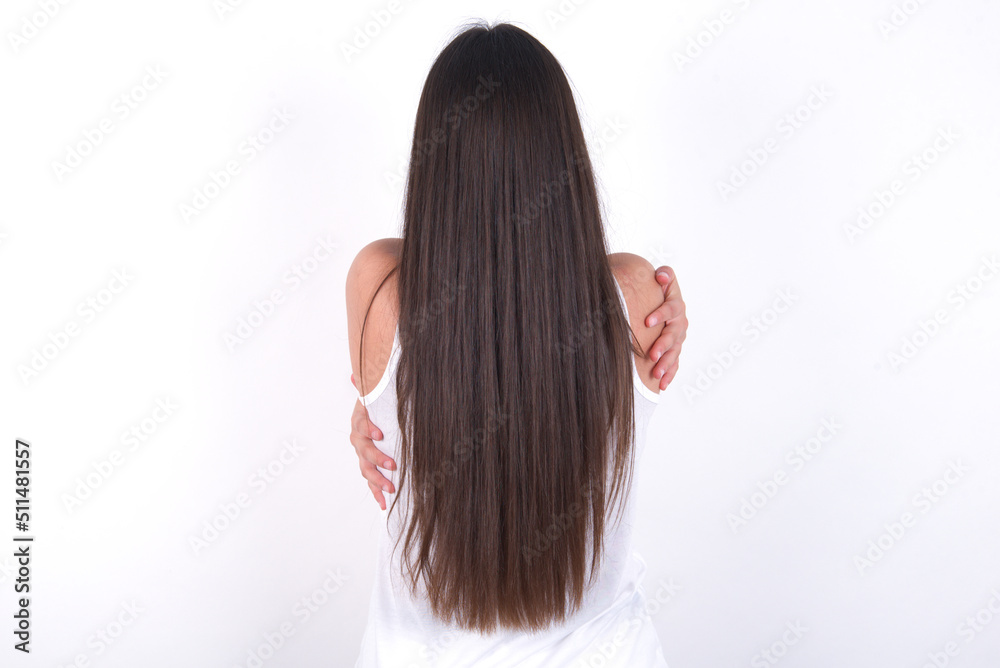 Young beautiful brunette woman wearing white top over white wall hugging oneself happy and positive from backwards. Self love and self care. Young beautiful brunette woman wearing white top over white wall hugging oneself happy and positive from backwards. Self love and self care.