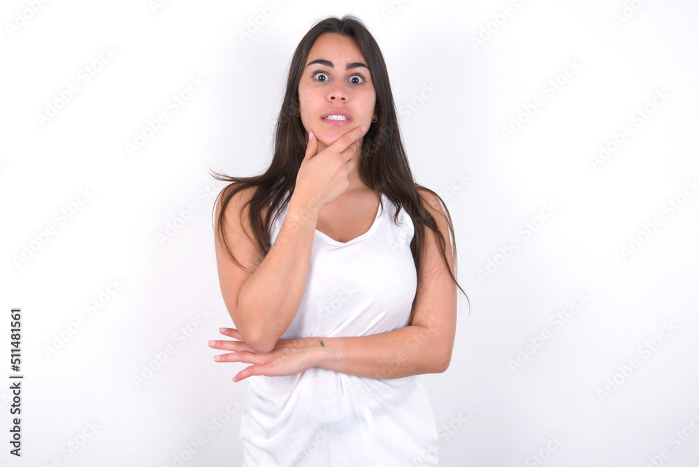 Young beautiful brunette woman wearing white top over white wall covering mouth with hands scared from something or someone bitting nails Young beautiful brunette woman wearing white top over white wall covering mouth with hands scared from something or someone bitting nails