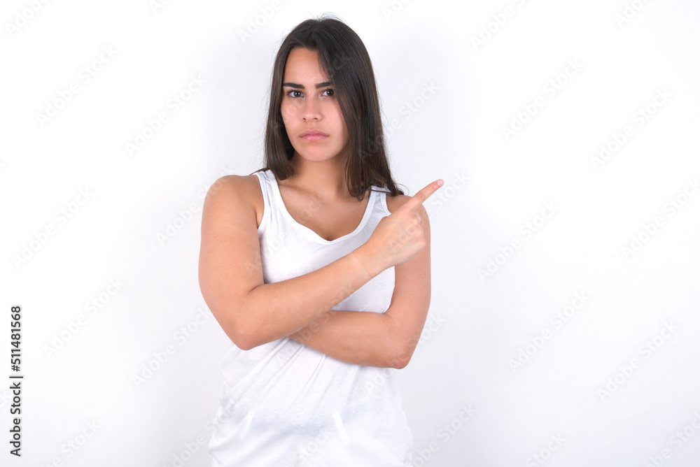 Young beautiful brunette woman wearing white top over white wall smiling broadly at camera, pointing fingers away, showing something interesting and exciting. Young beautiful brunette woman wearing white top over white wall smiling broadly at camera, pointing fingers away, showing something interesting and exciting.