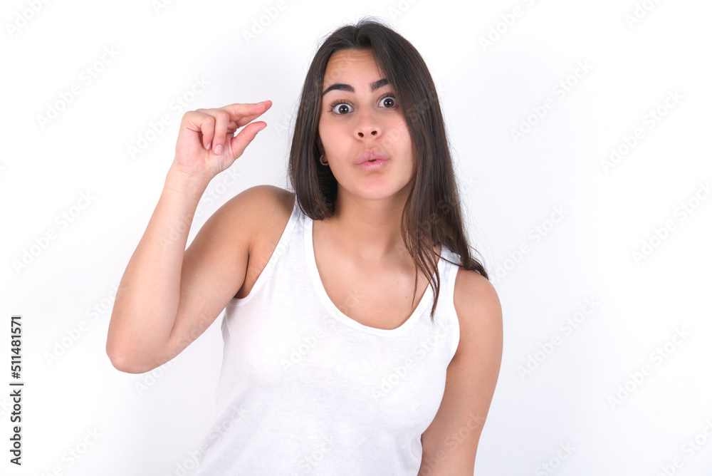 Shocked Young beautiful brunette woman wearing white top over white wall shows something little with hands, demonstrates size, opens mouth from surprise. Measurement concept. Shocked Young beautiful brunette woman wearing white top over white wall shows something little with hands, demonstrates size, opens mouth from surprise. Measurement concept.