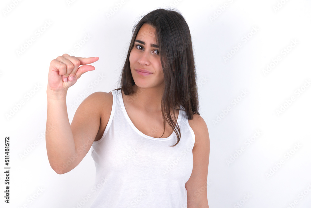 Upset Young beautiful brunette woman wearing white top over white wall shapes little gesture with hand demonstrates something very tiny small size. Not very much Upset Young beautiful brunette woman wearing white top over white wall shapes little gesture with hand demonstrates something very tiny small size. Not very much