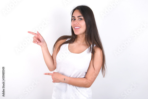 Young beautiful brunette woman wearing white top over white wall points aside with surprised expression with mouth opened, shows something amazing. Advertisement concept.