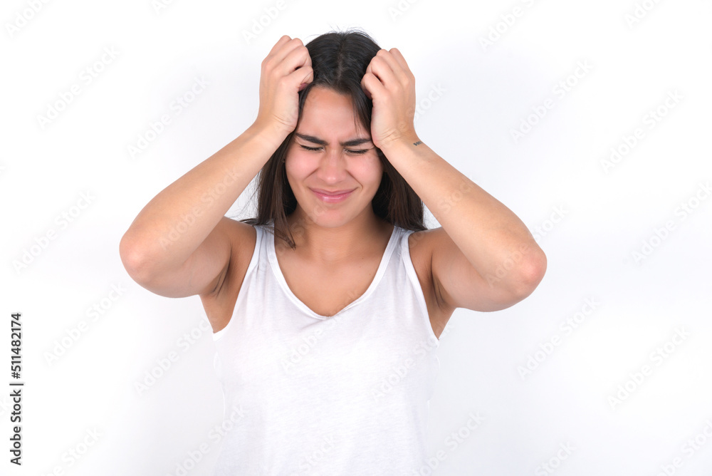 Young beautiful brunette woman wearing white top over white wall holding head with hands, suffering from severe headache, pressing fingers to temples Young beautiful brunette woman wearing white top over white wall holding head with hands, suffering from severe headache, pressing fingers to temples