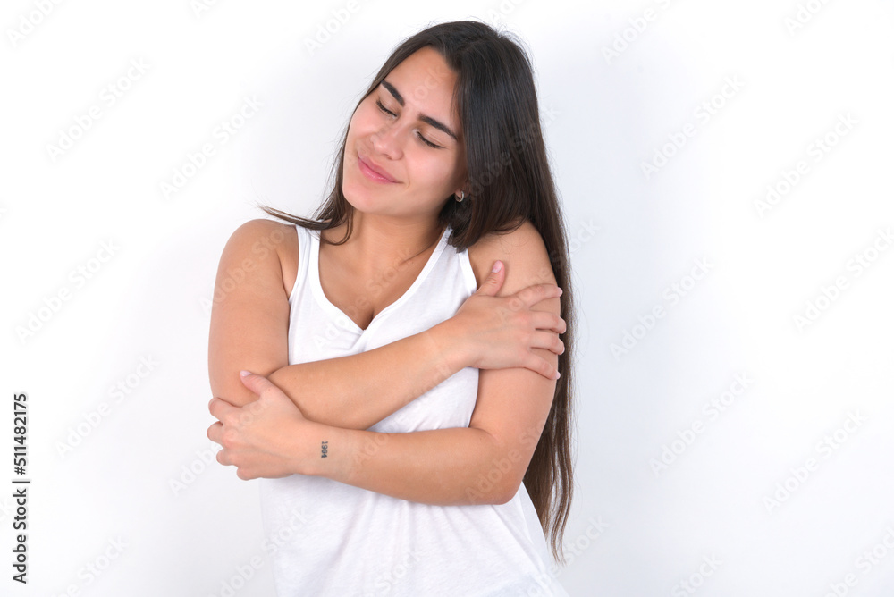 Young beautiful brunette woman wearing white top over white wall. Hugging oneself happy and positive, smiling confident. Self love and self care. Young beautiful brunette woman wearing white top over white wall. Hugging oneself happy and positive, smiling confident. Self love and self care.
