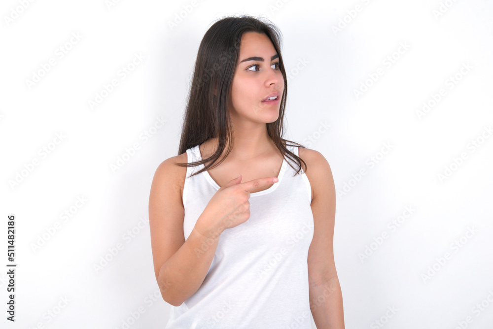 Happy cheerful smiling Young beautiful brunette woman wearing white top over white wall looking and pointing aside with hand. Copy space and advertisement concept. Happy cheerful smiling Young beautiful brunette woman wearing white top over white wall looking and pointing aside with hand. Copy space and advertisement concept.