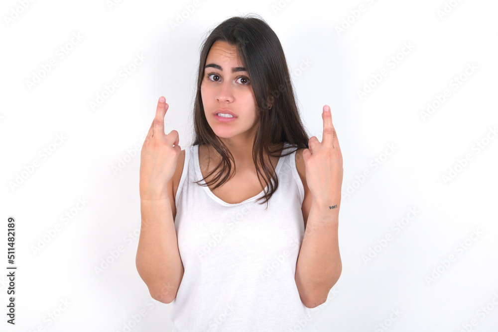 Young beautiful brunette woman wearing white top over white wall holding fingers crossed with worried expression hoping boss didn't noticed mistakes at work. Young beautiful brunette woman wearing white top over white wall holding fingers crossed with worried expression hoping boss didn't noticed mistakes at work.