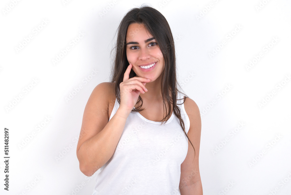 Carefree successful Young beautiful brunette woman wearing white top over white wall touching jawline gazing camera tilting head grinning white teeth delighted. Dental care concept. Carefree successful Young beautiful brunette woman wearing white top over white wall touching jawline gazing camera tilting head grinning white teeth delighted. Dental care concept.